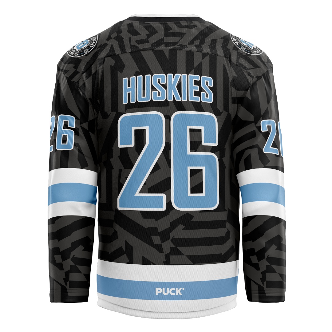 Back view of the Haringey Huskies 26/27 Away Jersey (NAMED) from the Haringey Huskies Store, featuring light blue and white accents, HUSKIES and the number 26 in large light blue font, and a pattern of dark geometric shapes. The word PUCK appears near the bottom. This jersey can be customized with your own name and number.