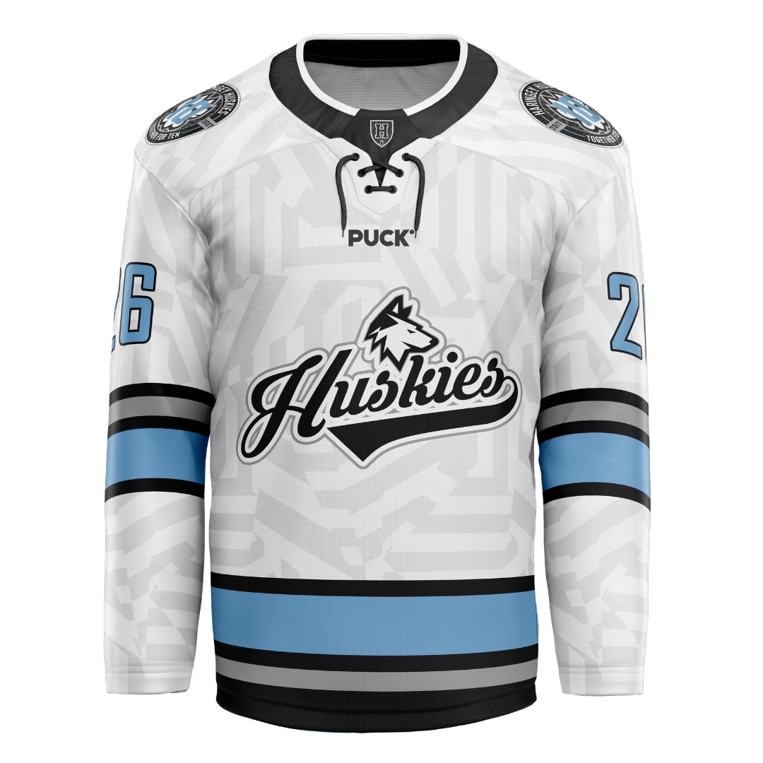 The Haringey Huskies 26/27 Home Jersey (NAMED) from the Haringey Huskies Store showcases a crisp white design accented with light blue and black stripes. Bold "Huskies" text and the iconic husky dog logo feature prominently on the chest, while the number 26 appears on both sleeves and circular patches decorate each shoulder. As a fan replica jersey, it can also be personalized with your choice of custom name and number for an extra-special touch.